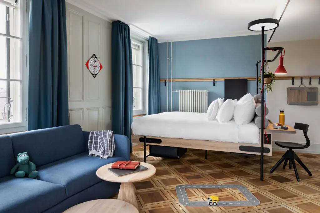 Bed and dark blue sofa with table on wooden floor in a bright room with window and decorative elements.