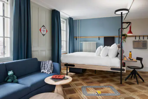 Bett und dunkelblaues Sofa mit Tisch auf Holzboden in hellem Raum mit Fenster und dekorativen Elementen.