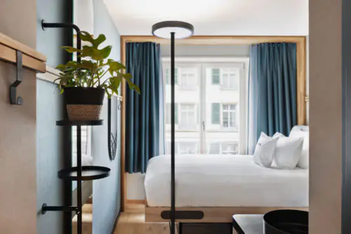 Helles Hotelzimmer mit Bett, weißer Bettwäsche und blauen Vorhängen, vorne Stehlampe und Pflanze.