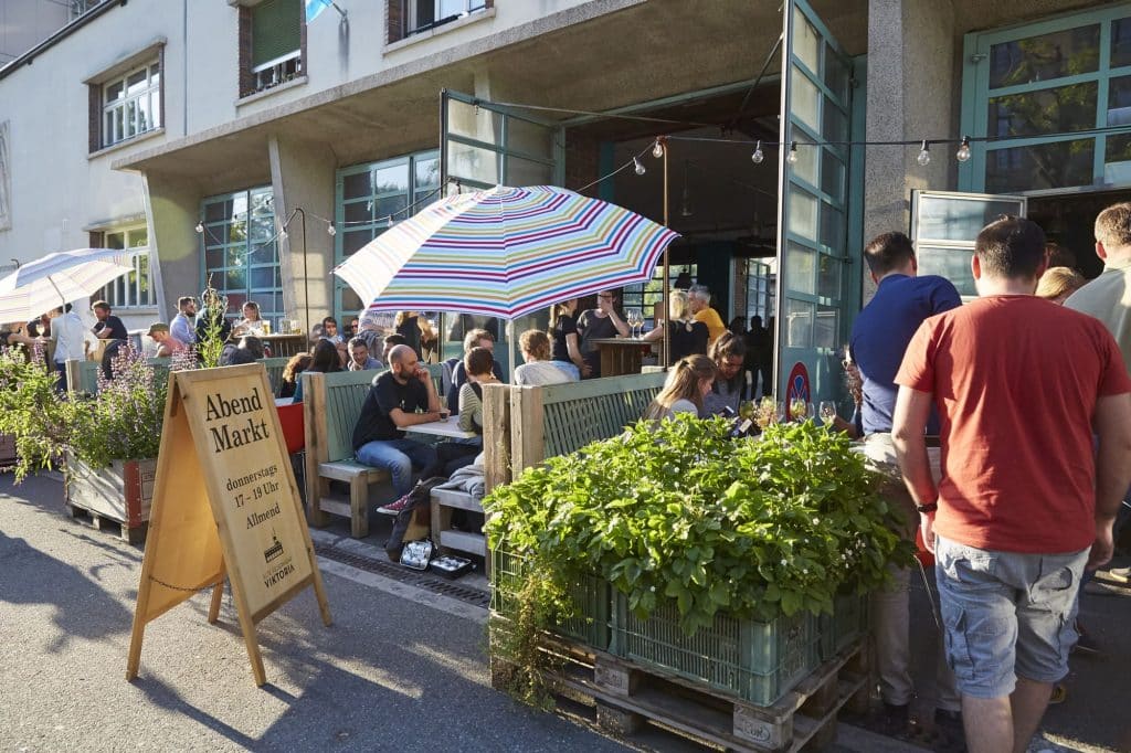 Belebter Außenbereich mit Menschen an Tischen, Sonnenschirm und Schild für Abendmarkt, entspannte Atmosphäre im urbanen Umfeld.