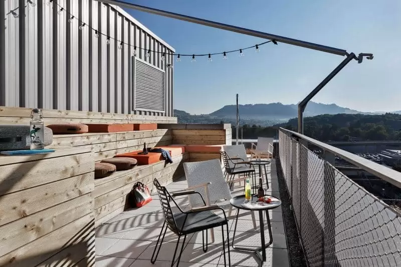 Rooftop terrace with tables, chairs, benches and string lights, with city and mountain views.