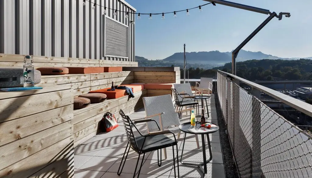 Rooftop-Terrasse mit Tischen, Stühlen, Bänken und Lichterkette, Blick auf Stadt und Berge im Hintergrund.