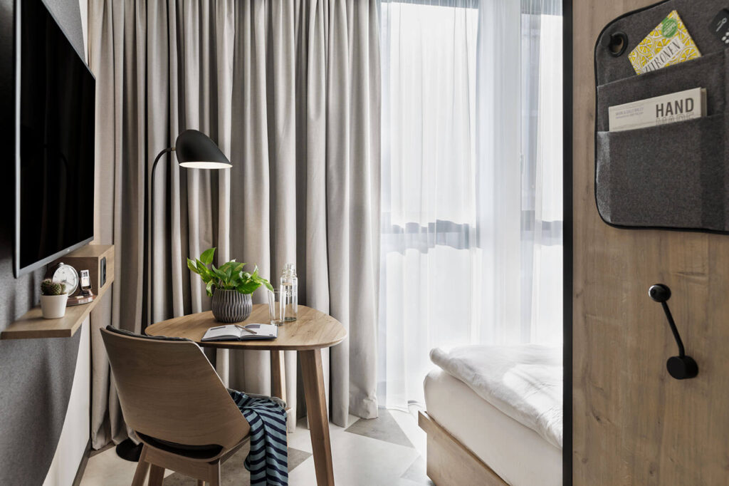 Helles Studio mit TV, Schreibtisch, Pflanze und Stuhl am Fenster, daneben Bettende und Schrank mit Haken.