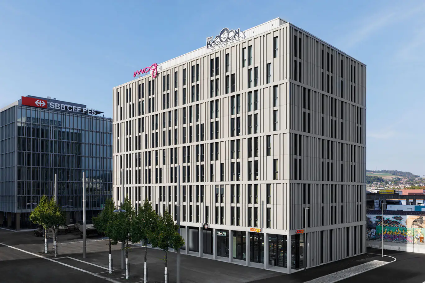 Außenansicht modernes Gebäude mit Moxy und Stay KooooK Logo, klare Fassadenstruktur, umgeben von Bäumen.