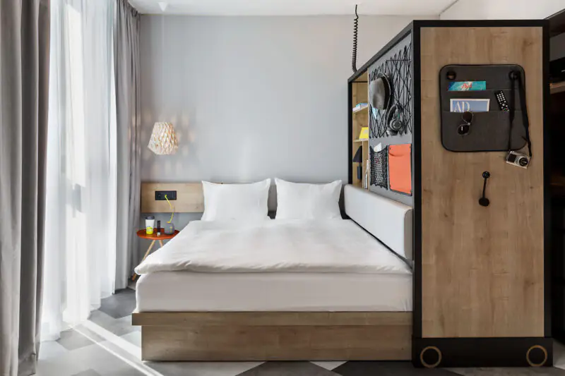 Studio with large window, double bed, bedside table, light wood interior and integrated storage.