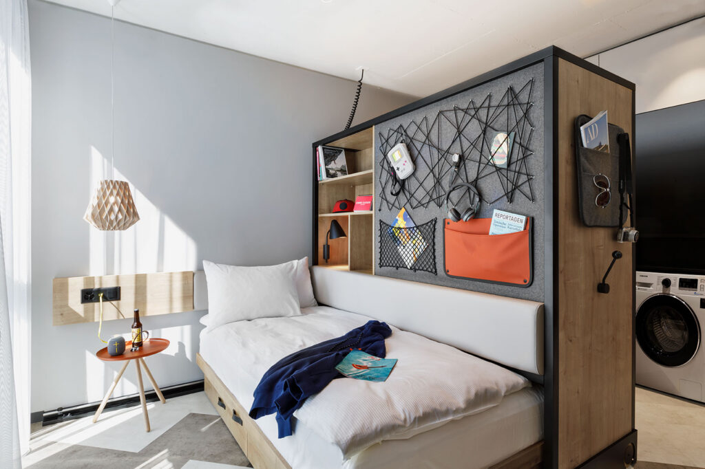 Studio mit Bett, Nachttisch, Lampe, Jacke und Heft, mit Ablageelement und Waschmaschine im Hintergrund.