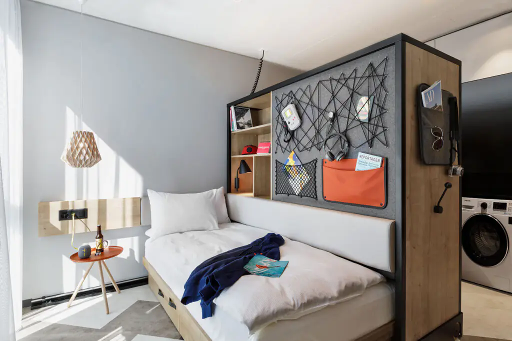 Studio with bed, bedside table, lamp, jacket and magazine, with storage element and washing machine behind.