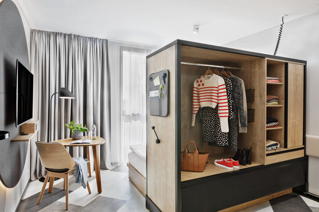 Studio mit multifunktionalem Möbel: offenes Verschiebeelement mit Garderobe, Sitzbereich und Fensterfront.