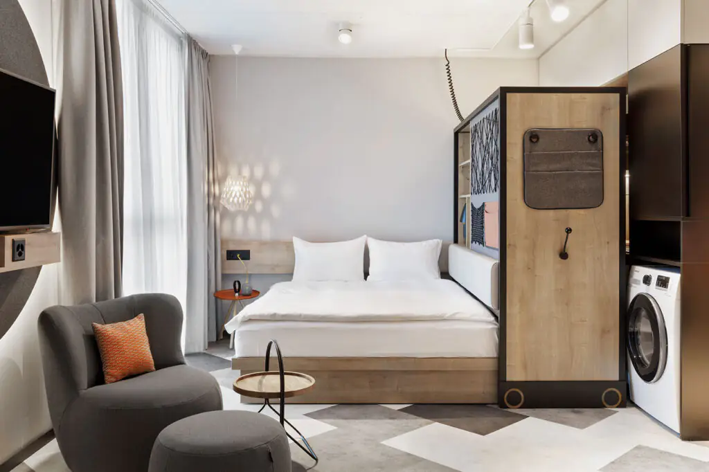Bett mit heller Bettwäsche, daneben offener Kleiderschrank, Waschmaschine und gemütliche Sitzecke.