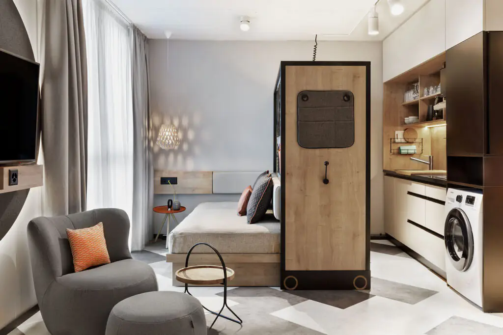 Bett mit Kissen, daneben offener Kleiderschrank, Waschmaschine und Sitzecke – moderne Raumnutzung.