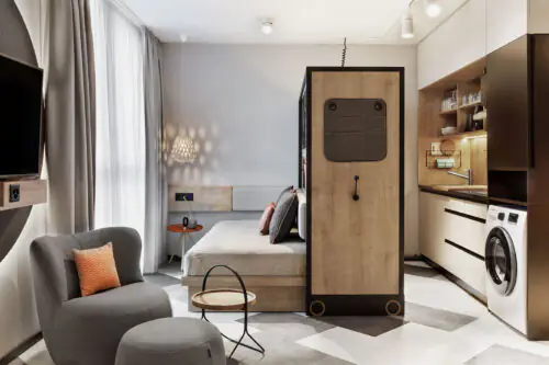 Bett mit Kissen, daneben offener Kleiderschrank, Waschmaschine und Sitzecke – moderne Raumnutzung.