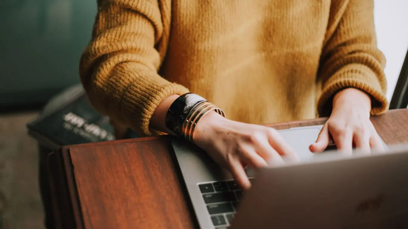 A person is working on a laptop at a wooden table and is wearing a mustard-yellow sweater.