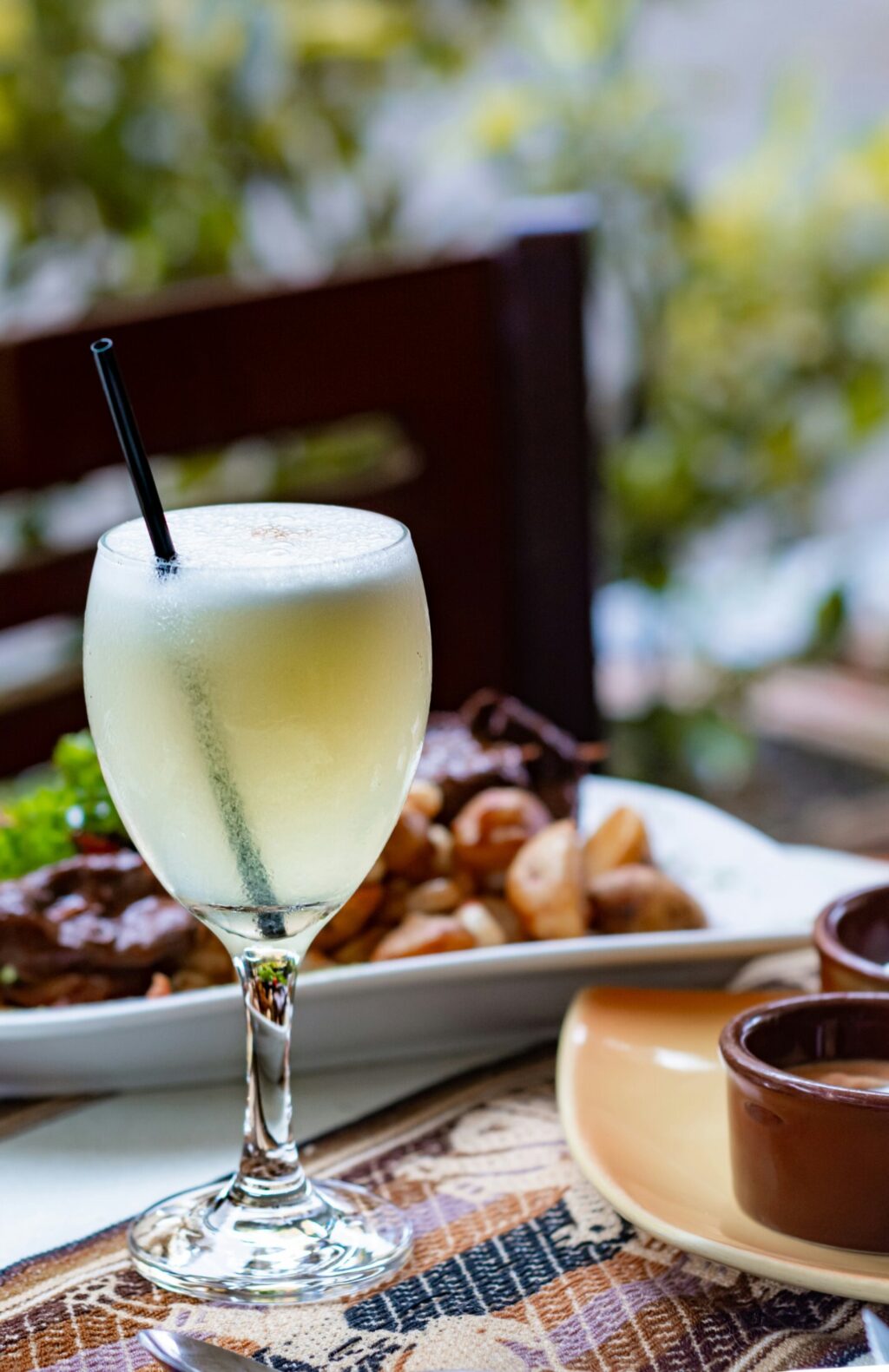Erfrischendes Getränk in Weinglas mit Strohhalm auf Tisch, im Hintergrund ein Teller mit Essen – sommerliche Restaurantatmosphäre.