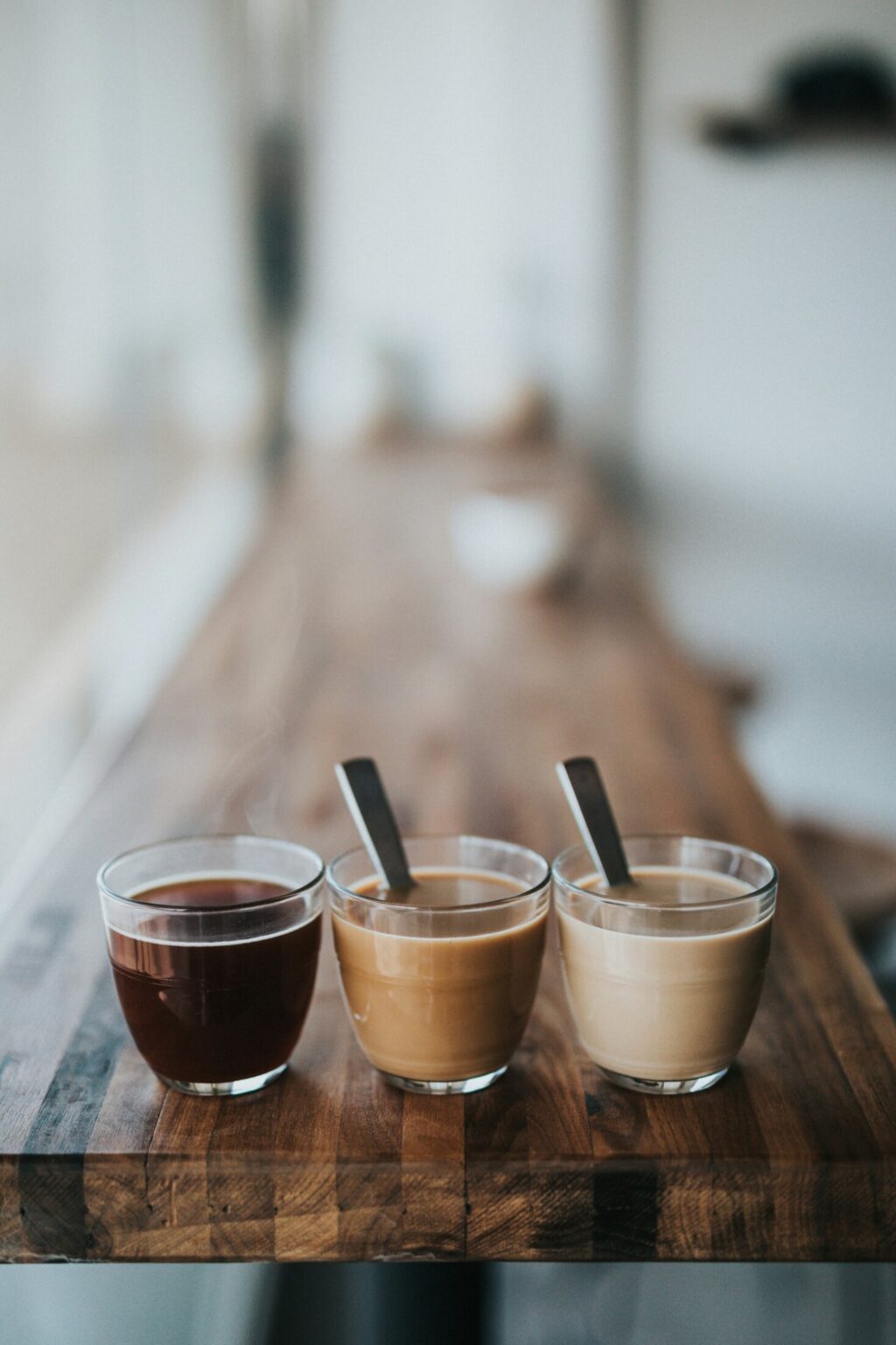 Drei Gläser Kaffee in unterschiedlichen Farben auf Holztisch – von schwarz bis milchig, mit Löffeln.