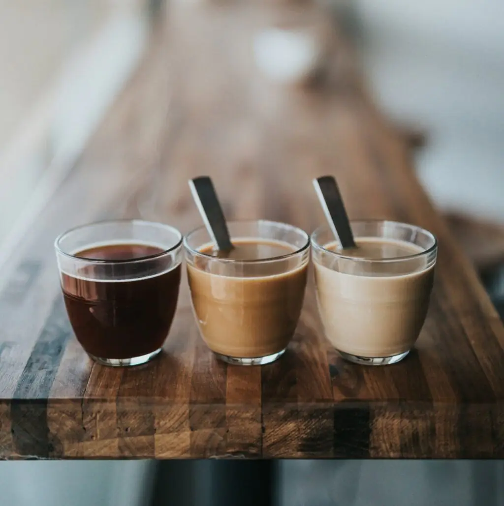 Drei Gläser Kaffee in unterschiedlichen Farben auf Holztisch – von schwarz bis milchig, mit Löffeln.