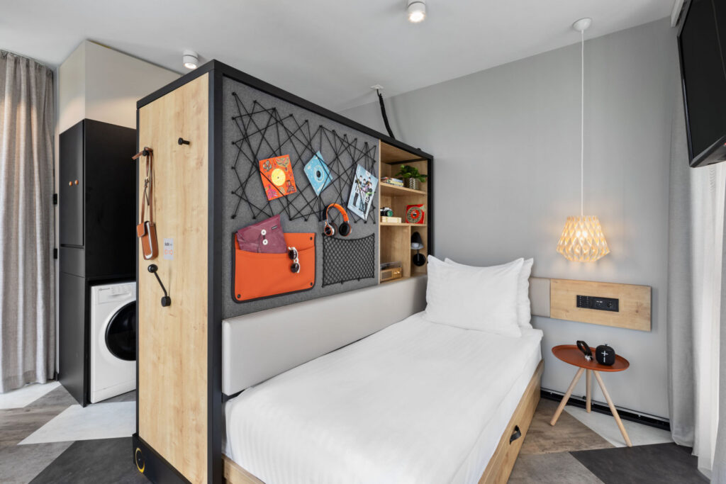 Studio mit Verschiebeelement hinter dem Bett mit Karten und Taschen, im Hintergrund Waschmaschine sichtbar.
