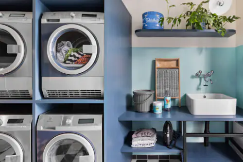 Laundry area with washing machines, dryers, shelf with plants and a sink in a modern space.