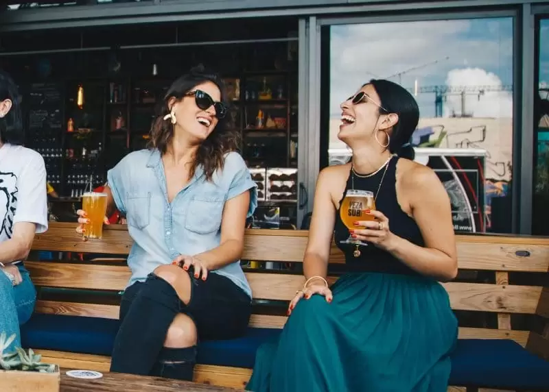 Zwei Frauen sitzen lachend auf einer Bank vor einer Bar und halten jeweils ein Getränk in der Hand.