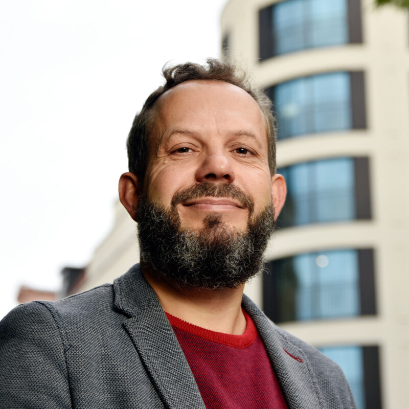 Man in business outfit in front of a building with round tower, shot from the side.