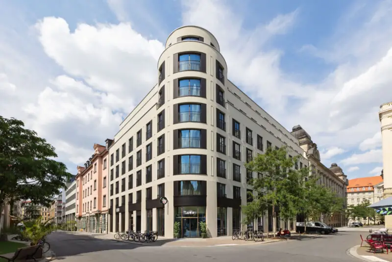 Frontalansicht eines modernen Stadthotels mit heller Fassade, Rundturm und großen Fenstern.
