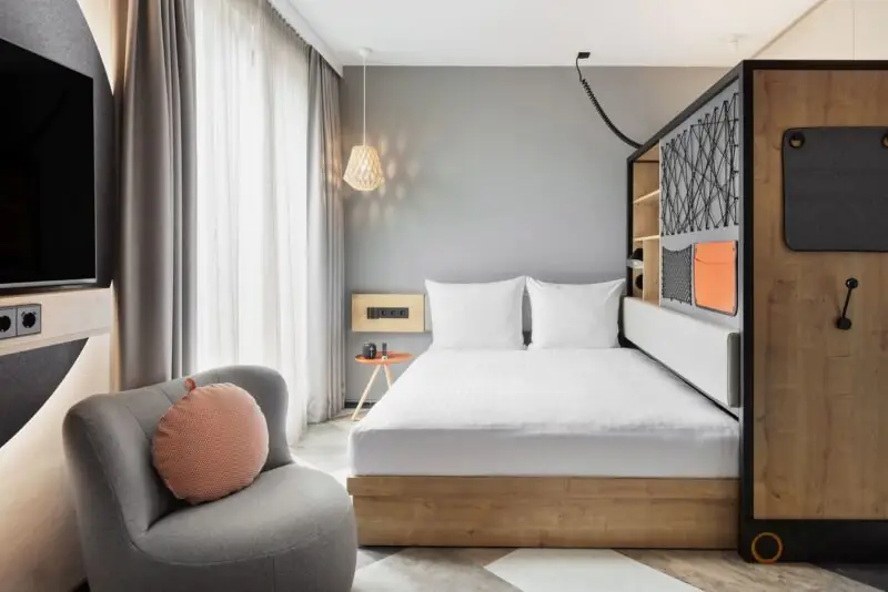 Helles Hotelzimmer mit Doppelbett, Sessel, TV und Stauraum – minimalistisch und einladend.