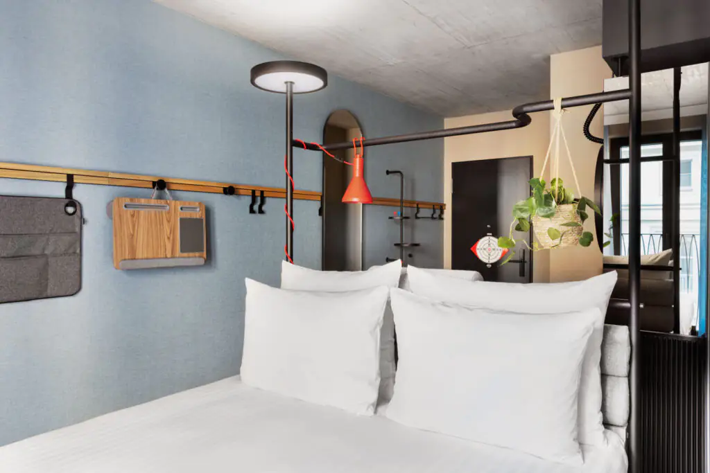 Double bed with white bedding against a light blue wall, reading lamp, plants and coat rack.