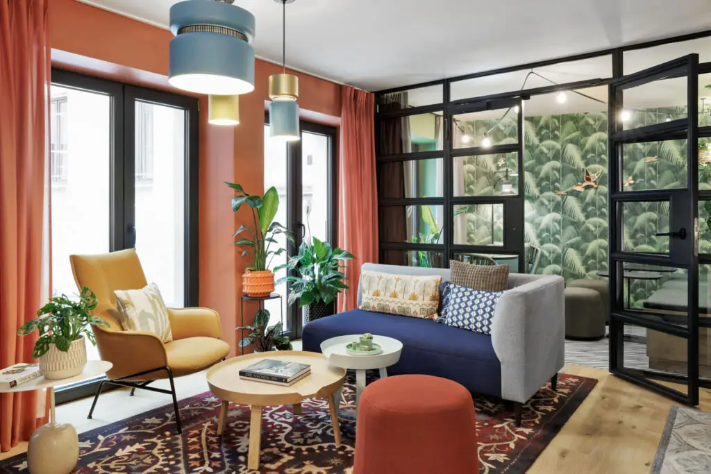 Farbenfroher Loungebereich mit Sofa, Sesseln, großen Fenstern und Blick ins Schlafzimmer mit Palmentapete.