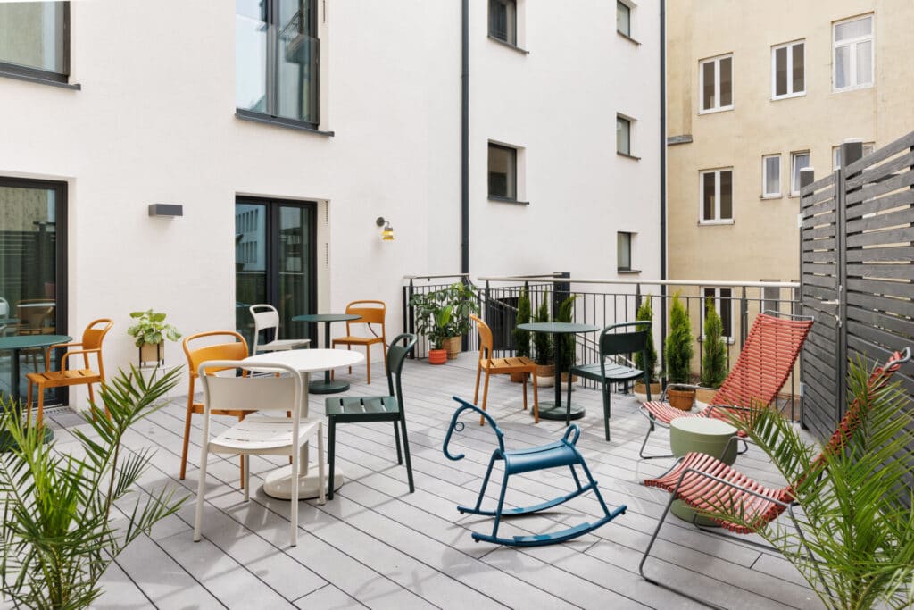 Dachterrasse mit bunten Stühlen und Tischen vor Wohngebäuden in entspannter Atmosphäre.