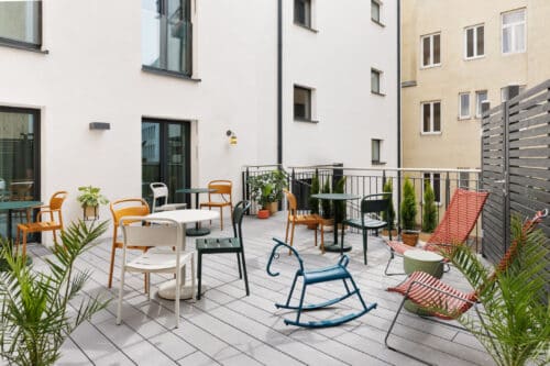 Dachterrasse mit bunten Stühlen und Tischen vor Wohngebäuden in entspannter Atmosphäre.
