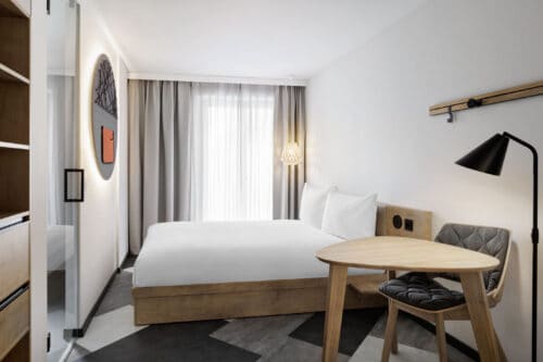Helles Hotelzimmer mit großem Fenster, Schreibtisch aus Holz und minimalistischer Einrichtung.