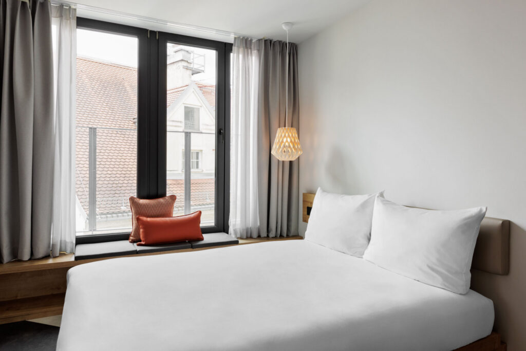 Bright room with double bed and large window, window nook with cushions in warm tones.