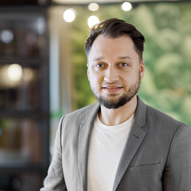 Portrait of host Alex smiling in a grey blazer in front of a green wall with decorative lighting.