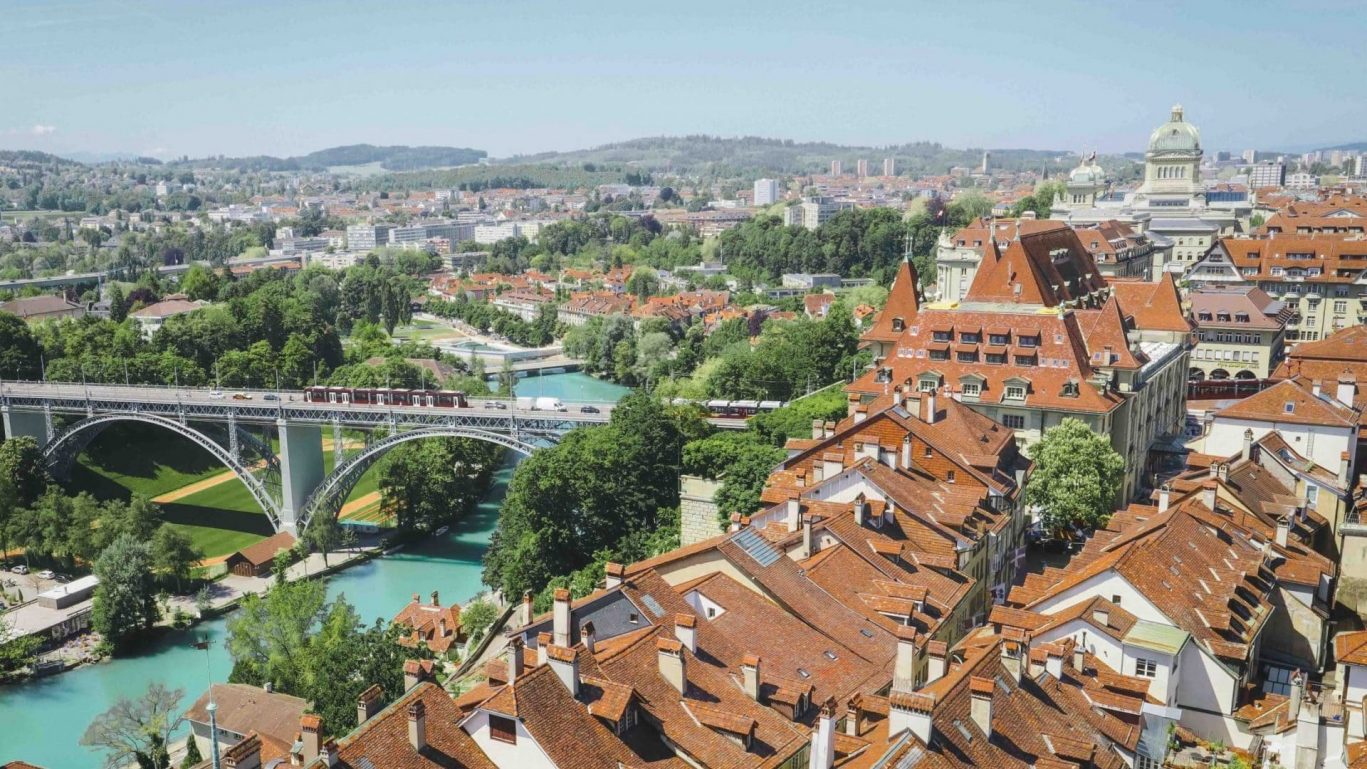 Luftaufnahme der Berner Altstadt mit roten Dächern, Aare und Brücke im Vordergrund.