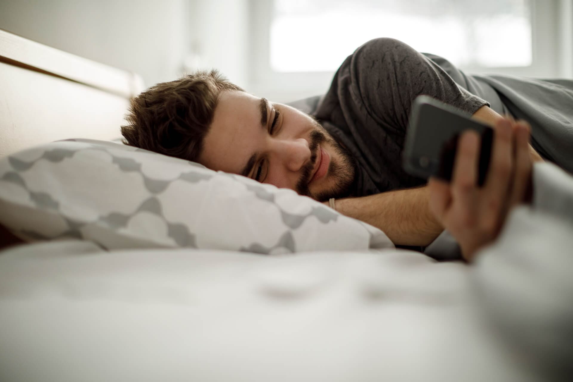 Entspannter Mann liegt im Bett und schaut lächelnd auf sein Smartphone.