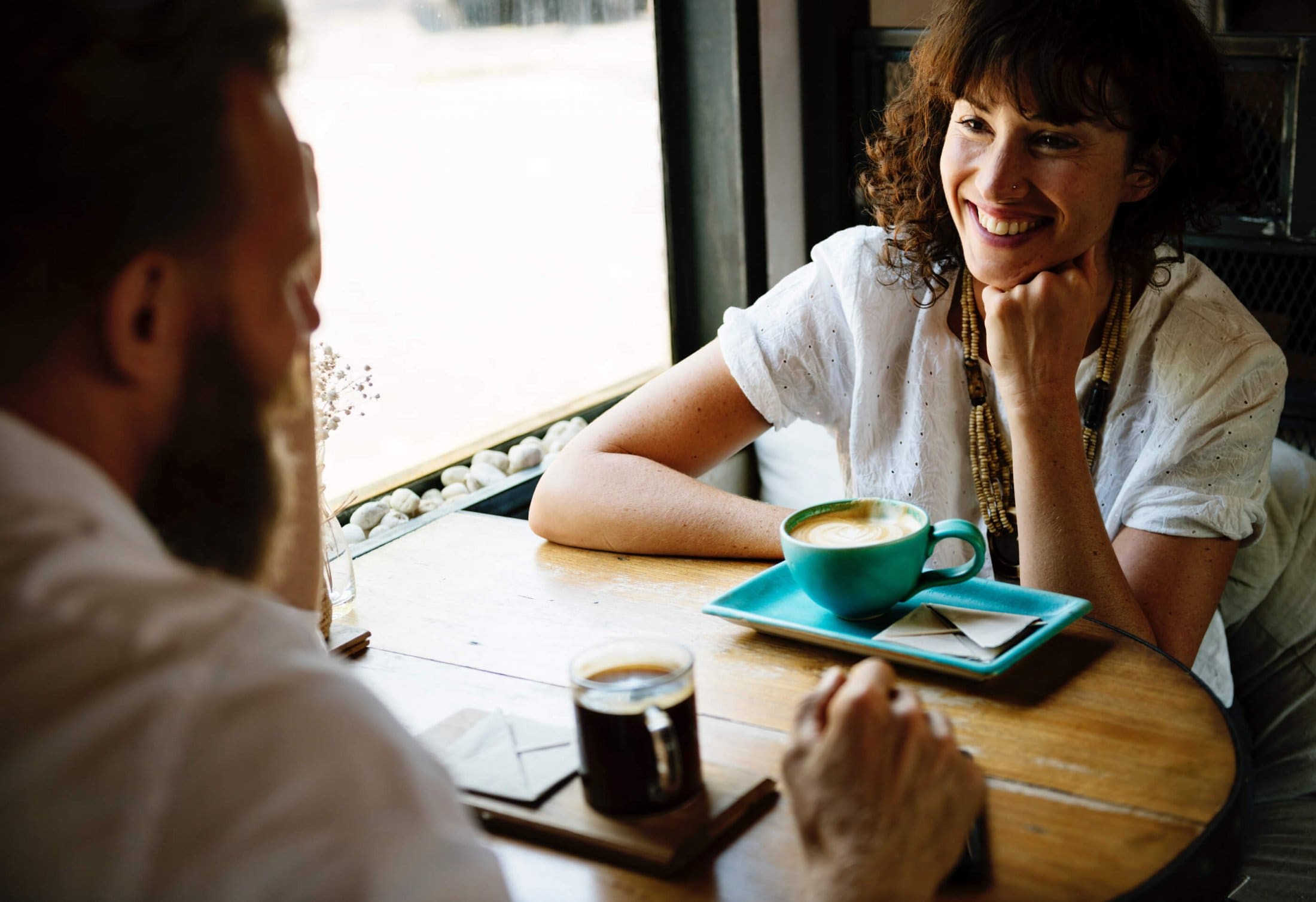 Two people sit in a café, drinking coffee and talking at a wooden table by the window.