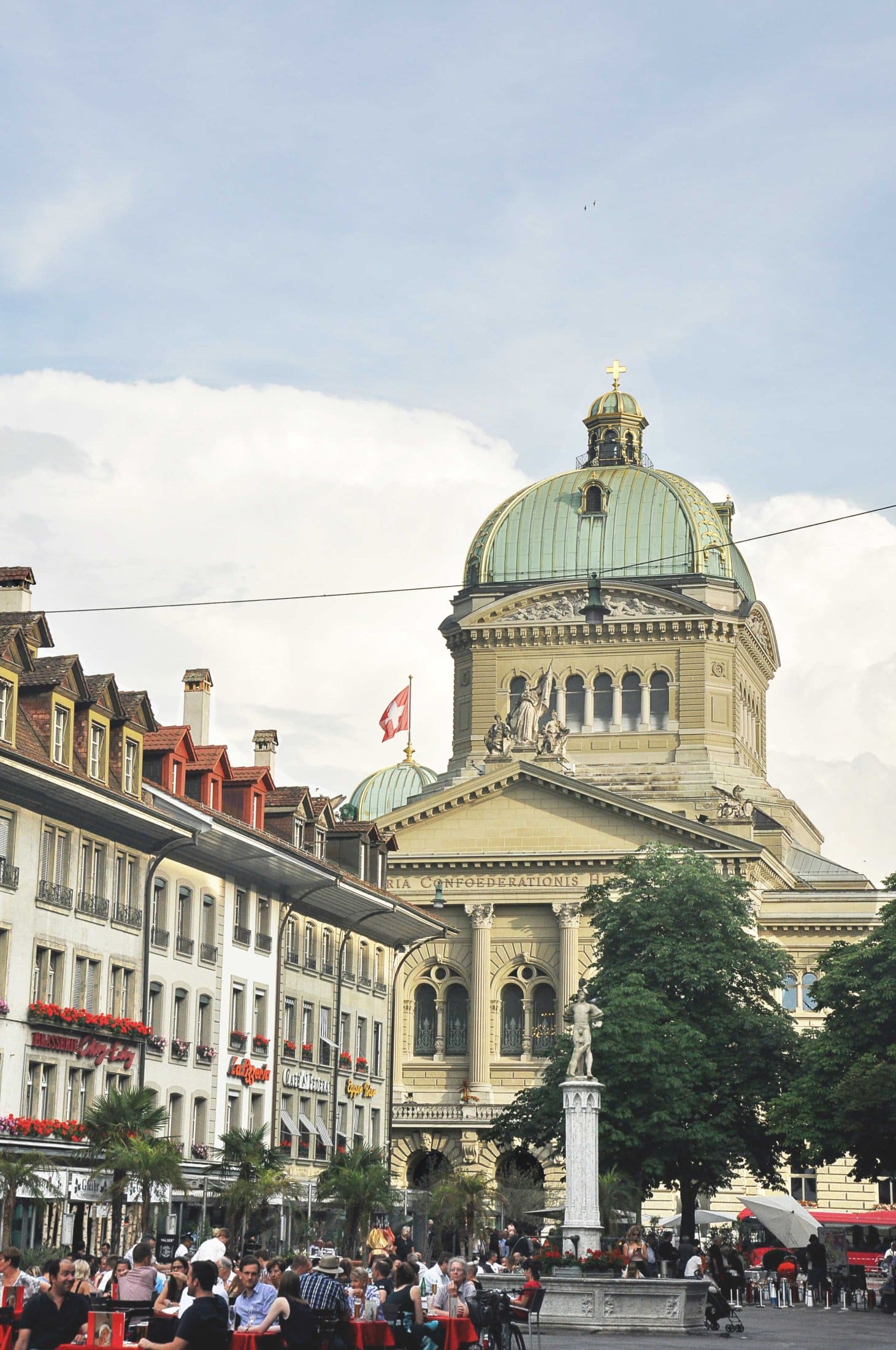 Federal Palace in Bern with lively square, cafés and historic architecture under a bright city sky.