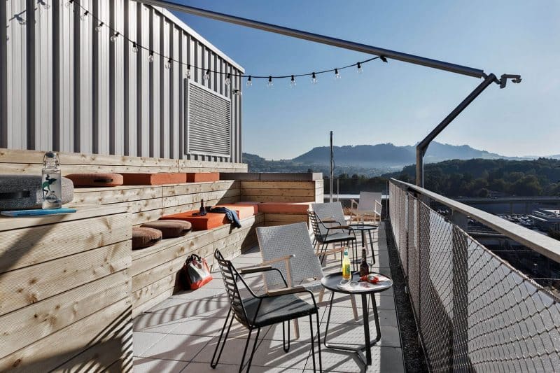 Rooftop-Terrasse mit Tischen, Stühlen, Bänken und Lichterkette, Blick auf Stadt und Berge im Hintergrund.
