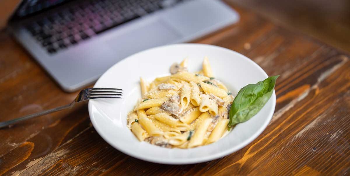 Teller mit cremiger Penne-Pasta, Pilzen und Parmesan auf Holztisch neben Laptop und Gabel angerichtet.