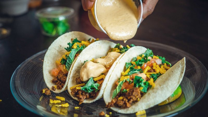 Three filled tacos are topped with creamy sauce, loaded with vegetables, cheese, and seasoned meat.