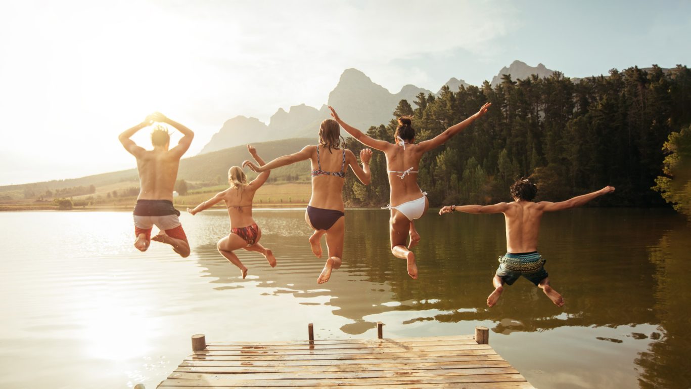 Gruppe von Freunden springt gemeinsam von einem Steg in einen See, umgeben von Bergen – vermittelt pure Lebensfreude, Freiheit und unbeschwerte Momente.