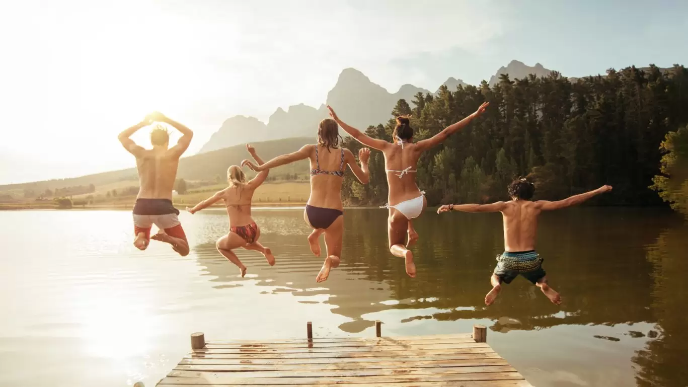 Gruppe von Freunden springt gemeinsam von einem Steg in einen See, umgeben von Bergen – vermittelt pure Lebensfreude, Freiheit und unbeschwerte Momente.