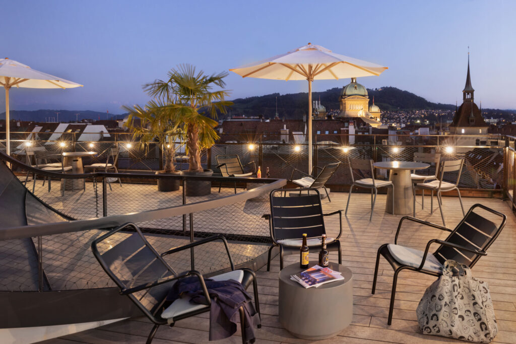 A rooftop terrace in Bern city centre with cosy seating areas, atmospheric lighting and views across the city to the Alps.