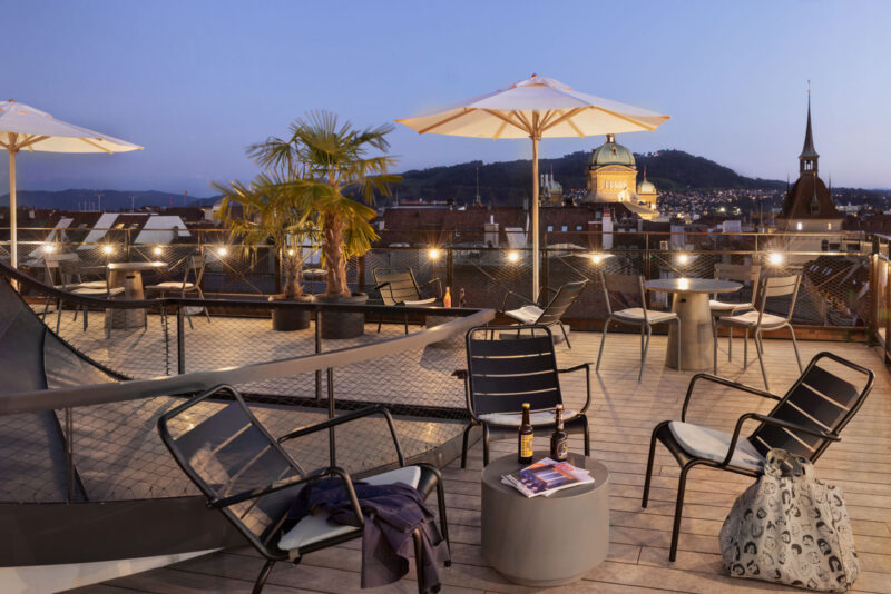 A rooftop terrace in Bern city centre with cosy seating areas, atmospheric lighting and views across the city to the Alps.