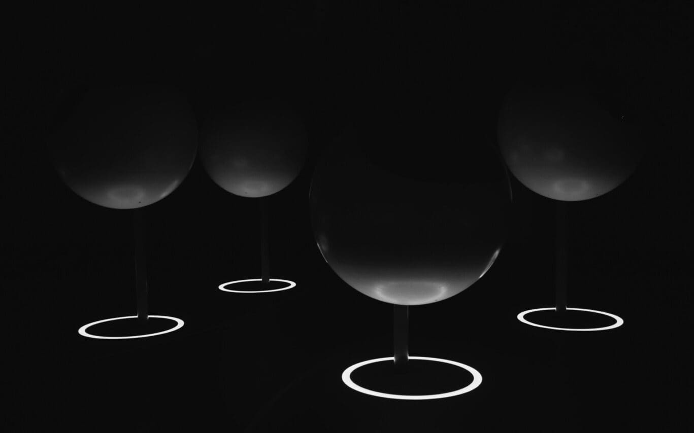 Mehrere Weingläser stehen im Dunkeln, nur von kreisförmigen Lichtakzenten am Fuß sanft beleuchtet – eine minimalistische, elegante und fast mystische Inszenierung.