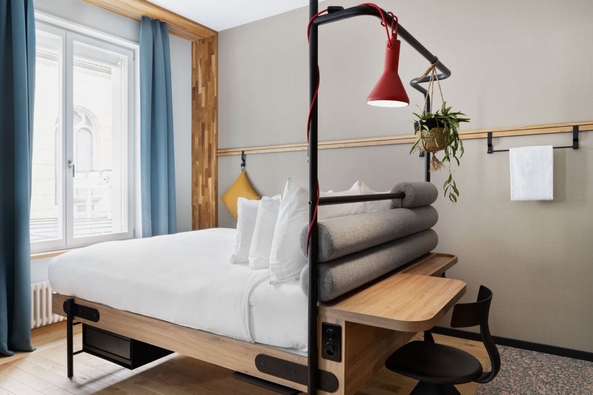 Detail eines Betts mit roter Lampe, Hängepflanze, kleinem Schreibtisch und Tageslicht von links.