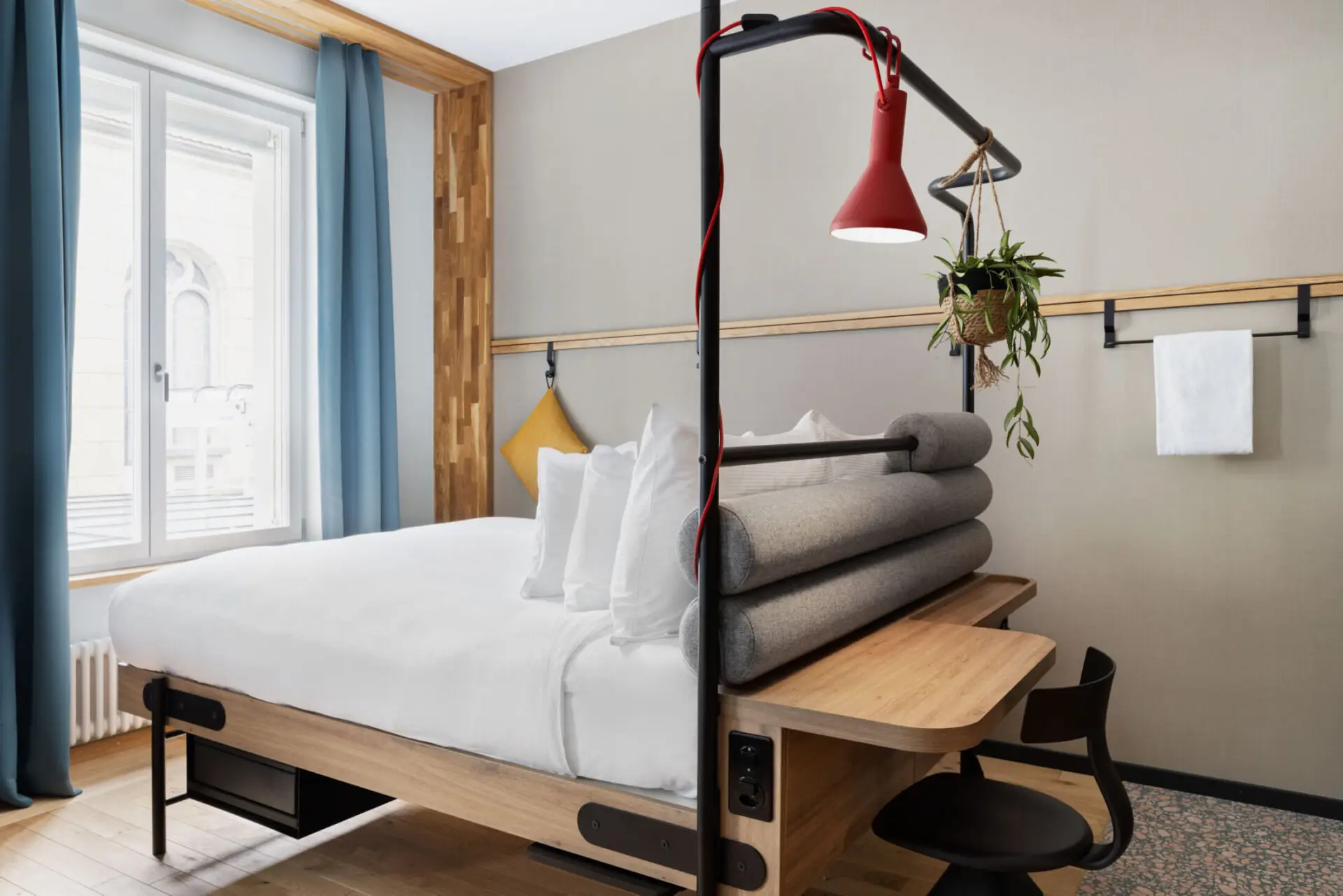 Detail eines Betts mit roter Lampe, Hängepflanze, kleinem Schreibtisch und Tageslicht von links.