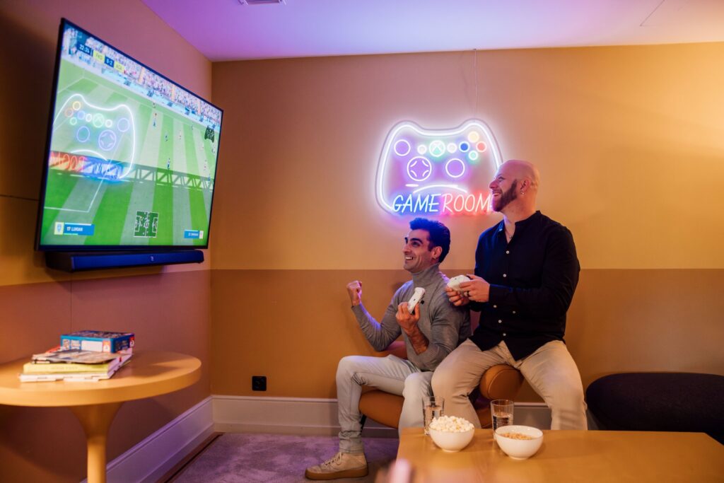 Zwei Männer spielen Fußball-Videospiel vor TV mit Controllern, Neon-Schild „Game Room“ im Hintergrund.
