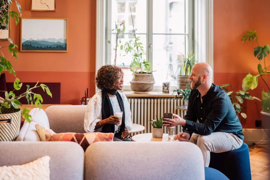 Two people sit on a sofa, drink coffee and chat in a bright lounge with plants and warm colors.