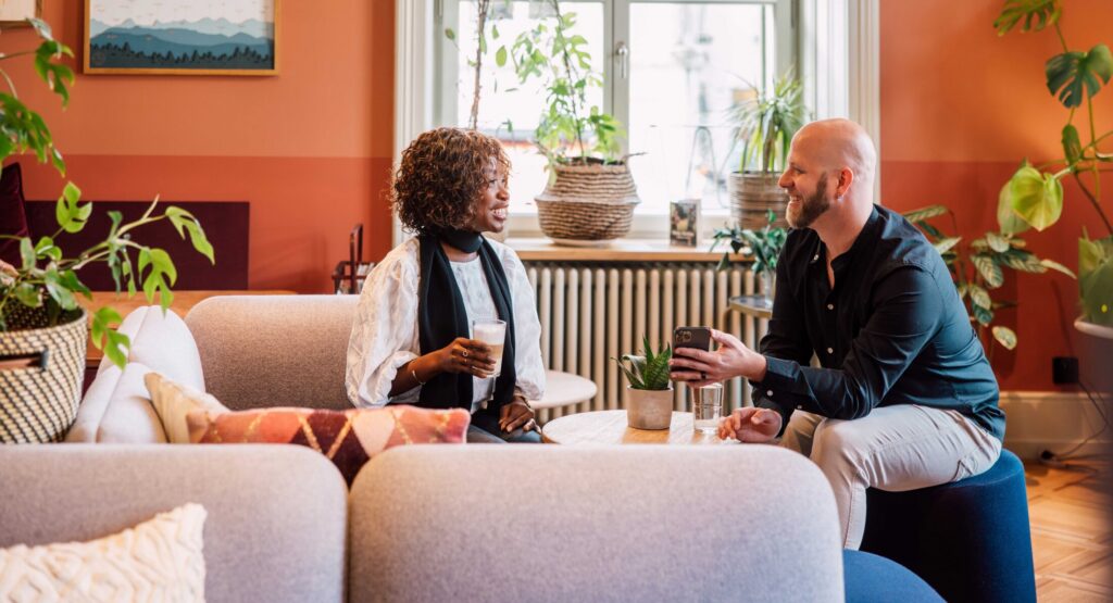 Zwei Personen sitzen auf Sofa, trinken Kaffee und unterhalten sich in heller Lounge mit Pflanzen und warmen Farben.