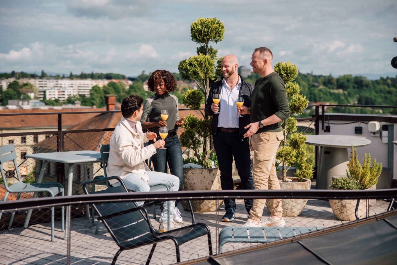 Vier Personen stehen auf der Rooftop Terrasse, trinken gemeinsam und genießen den Blick über Stadt und Umgebung.